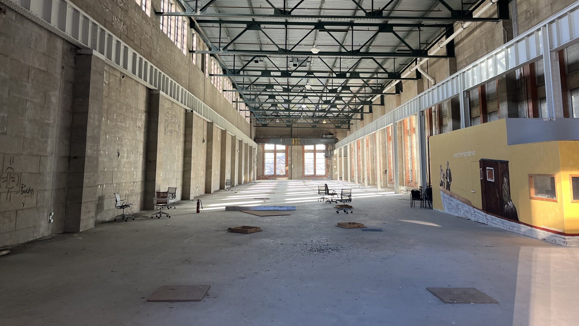 A current interior photo of the Haxall Canal Hydroelectric Power Plant. It is a massive room, with a new floor added by Cordish Companies, and with a yellow mural surrounding a power station. Natural light exposes the disarray which the building has fallen into before new ownership by Nordheimer Companies and Thalheimer Realty Partners transform the building into the Padel Plant.